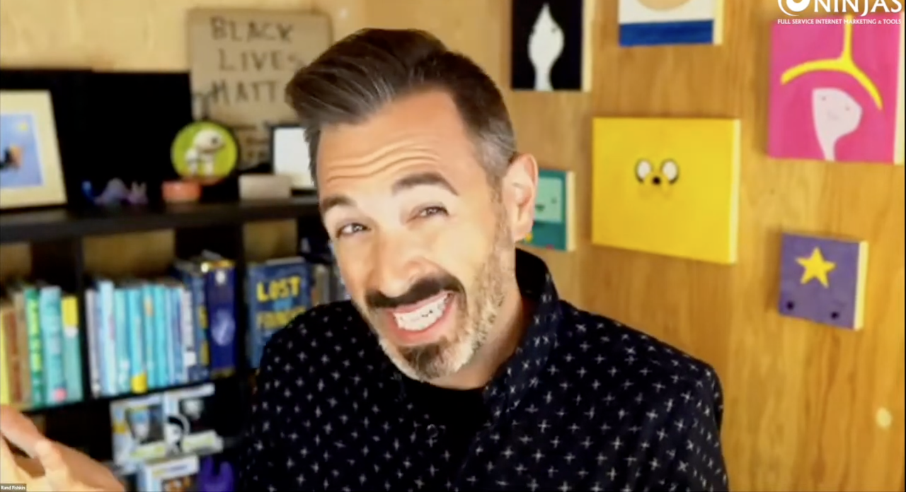 Rand Fishkin Man with dark hair and beard gestures while speaking on a video call about Product-Driven SEO, with colorful art and books behind him—a scene reminiscent of Rand Fishkin’s sessions for SEOs.