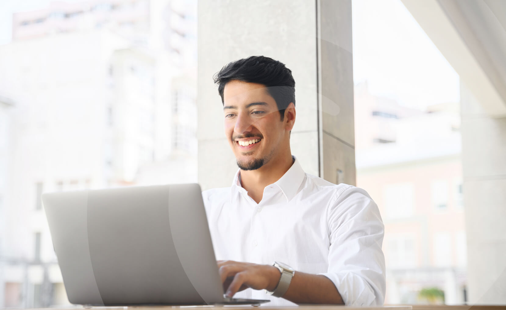 Man in a white shirt smiling while working on a laptop at a bright indoor location