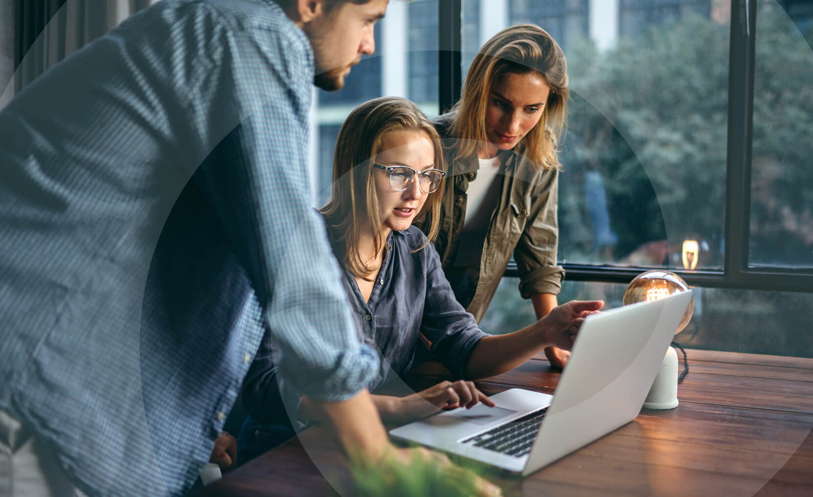 Three people working together at a desk, looking intently at a laptop near a large window