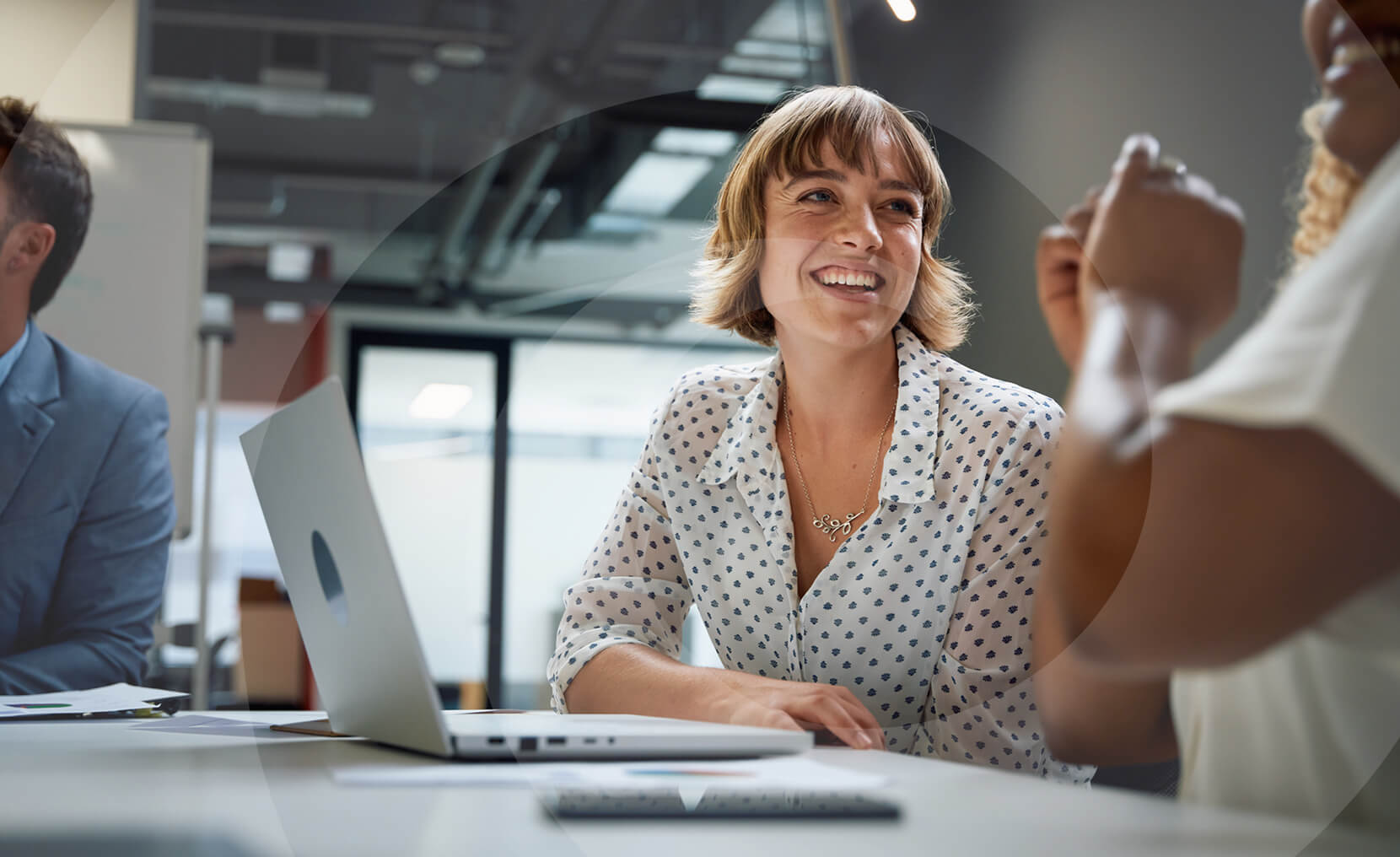 Smiling woman at a table with a laptop, engaged in conversation about social media optimization in a modern office setting.